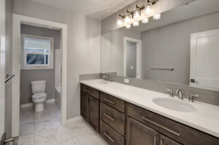 This is a modern bathroom with double sinks, wooden cabinets, a large mirror, tiled floor, and a separate toilet area with a window.