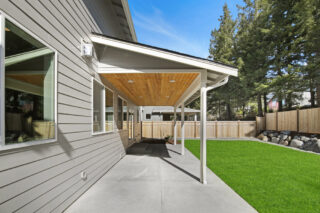 This image shows the side of a modern house with a wooden patio cover, green artificial lawn, tall trees, a fence, and a concrete walkway.