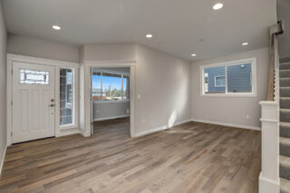 An empty room with hardwood floors, white walls, recessed lighting, a staircase, windows, and a white door with sidelights. Modern, clean, and bright.