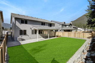 A two-story grey house with white trim, a well-manicured lawn, a wooden fence, and a mountainous background under a clear blue sky.