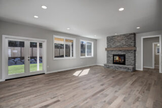 A spacious living room with a fireplace, wood flooring, and large windows that provide a view of a backyard with a wooden fence.