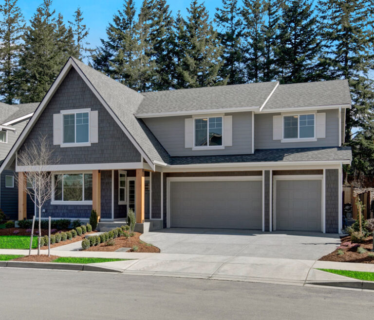  This image shows a two-story house with gray siding, white trim, a double garage, and a landscaped front yard, surrounded by tall evergreen trees under a clear sky. 