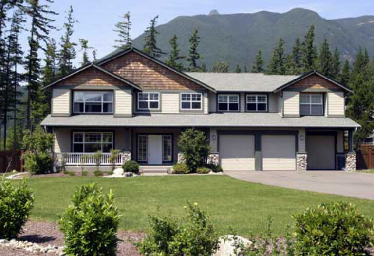  A two-story suburban house with a gabled roof, double garage, and a small porch. Landscaped with bushes, a lawn, and a backdrop of forested mountains. 