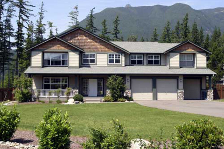 A two-story suburban house with a gabled roof, double garage, and a small porch. Landscaped with bushes, a lawn, and a backdrop of forested mountains.