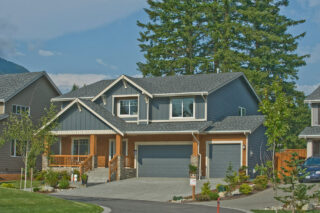 A modern two-story house with a gray exterior, double garage doors, and a wooden entrance porch, set against a backdrop of tall trees and mountains.