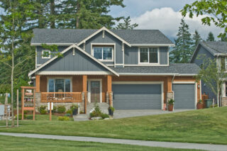 A two-story house with a grey exterior, white trim, wooden porch, double garage, and a well-manicured lawn. A sign indicates it as a model home.