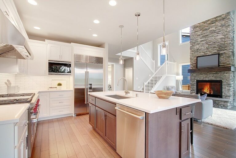 Modern kitchen with white cabinets, stainless steel appliances, a central island, pendant lights, hardwood floors, and a stone fireplace visible.