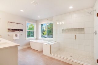 Modern bathroom with a freestanding tub, glass-enclosed shower, double windows, recessed lighting, abstract art, and neutral color palette.