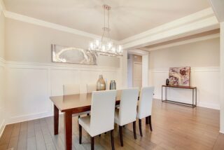 Elegant dining room featuring a wooden table, white chairs, chandelier, decorative art, wainscoting walls, and hardwood floors in a neutral-toned space.