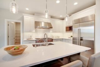 Modern kitchen interior with white cabinets, stainless steel appliances, pendant lights, a large island with sink, and a wooden fruit bowl.