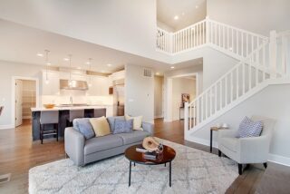 Modern, open-plan living space with a grey sofa, white kitchen, wooden floors, staircase leading to an upper level, and natural light.