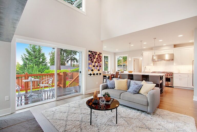 Modern living room with a gray sofa, round coffee table, large windows, white kitchen cabinets, and a view of a wooden deck outside.