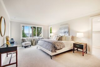 Bright, spacious bedroom with a large bed, two nightstands, lamps, a chair, decorative items, artwork, and a view of trees through windows.