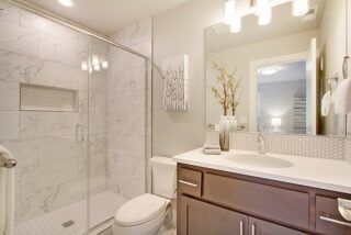 Modern bathroom with marble tiles, glass-door shower, wooden vanity, mirror with wall sconces, and decorative items on the counter. Clean, well-lit space.