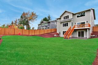 A two-story house with a wooden deck and stairs, a green lawn, and a wooden fence under a partly cloudy sky. No people are visible.