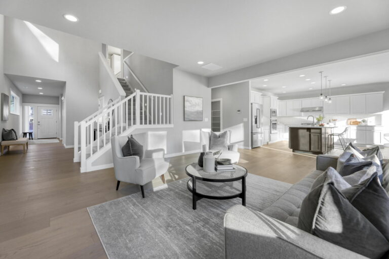 A modern, open-plan living space with a living area, kitchen, and staircase. Neutral colors, natural light, and contemporary furniture create a clean, airy atmosphere.