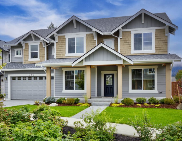  A two-story suburban house with beige and gray siding, a gabled roof, a front yard with green landscaping, and an attached garage. 