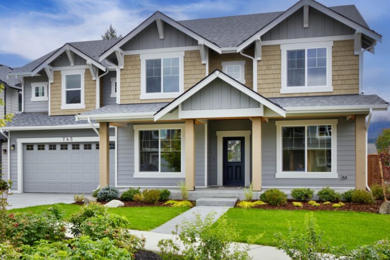 A two-story suburban house with beige and gray siding, a gabled roof, a front yard with green landscaping, and an attached garage.