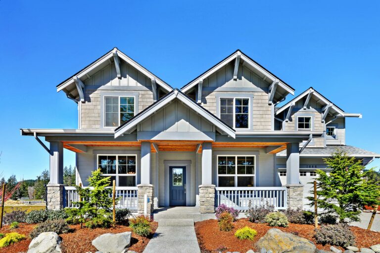 New two-story house with a symmetrical facade, prominent gables, stone column porch, landscaped front yard, under a clear blue sky.