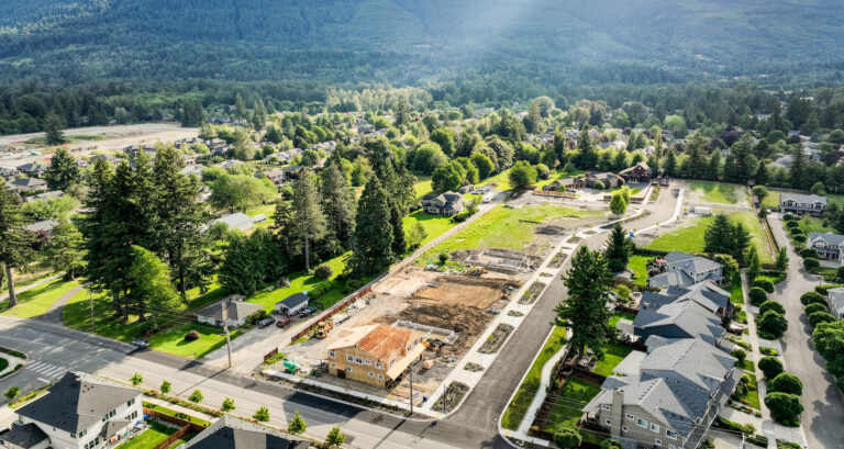 Aerial view of a suburban development with green trees, houses, and construction sites near a forested mountain range on a sunny day.