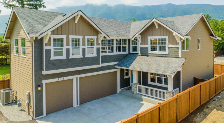 This is a two-story suburban house with a double garage, beige siding, white trim, and a small front porch, surrounded by a wooden fence, with mountains in the distance.