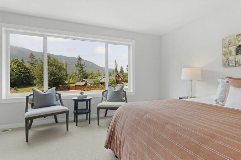 A clean, bright bedroom with a large bed, two chairs, a side table, and a lamp. Expansive windows offer views of mountains and greenery.