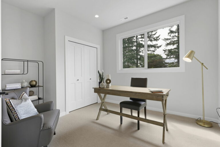 A serene home office with a wooden desk, chair, floor lamp, bookshelf, a comfortable armchair, and a window showcasing a view of pine trees.