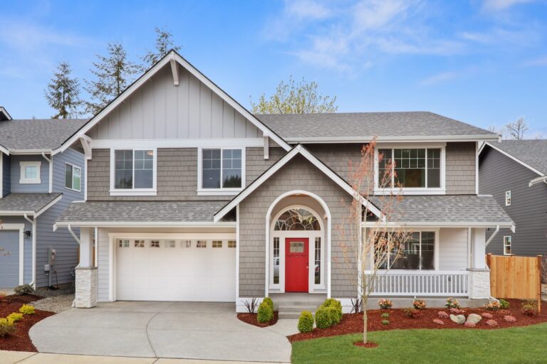 A two-story suburban house with gray siding, white trim, a red door, attached garage, landscaped front yard, and a clear blue sky.