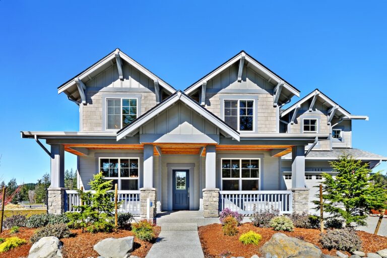  A craftsman style house features a gabled roof, a covered front porch with stone pillars, large windows, and beautiful landscaping under a blue sky. 