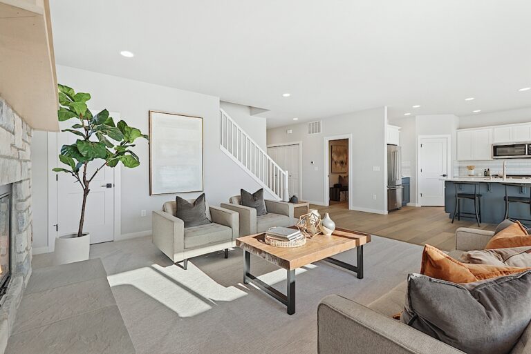 A bright, spacious living room with modern decor, stone fireplace, armchairs, wood coffee table, area rug, and an open kitchen in the background.