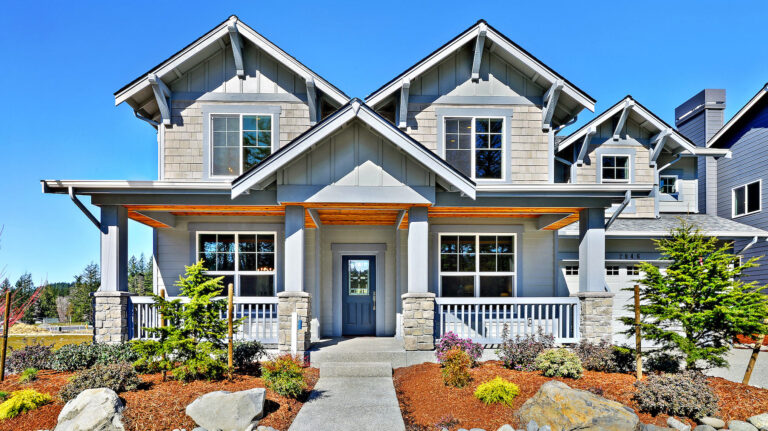  A two-story house with a gabled roof, stone column accents, covered porch, and landscaped garden under a clear blue sky. 