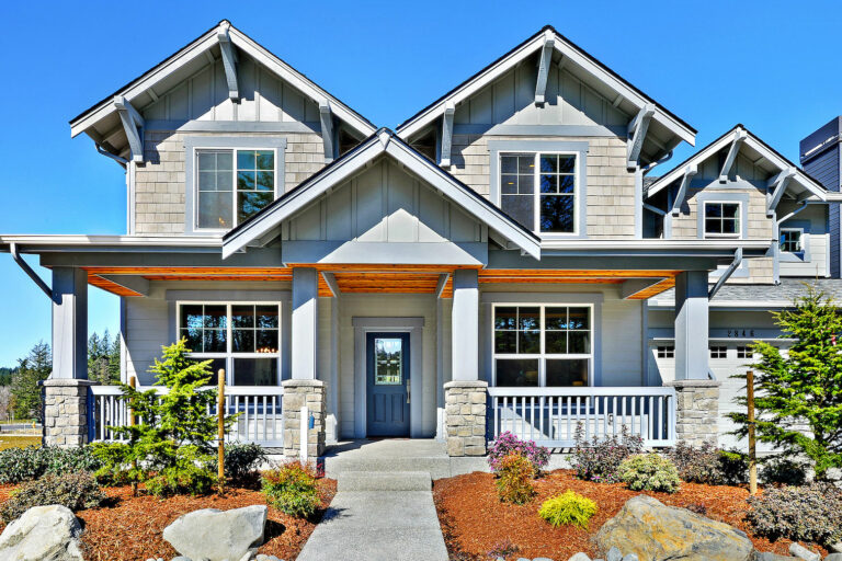 A two-story house with a gabled roof, stone column accents, covered porch, and landscaped garden under a clear blue sky.