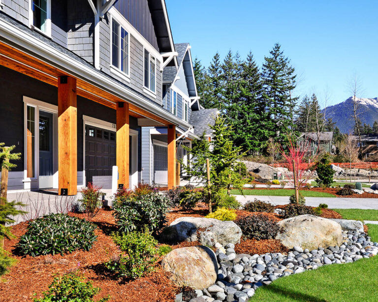 A suburban house with a gray and wooden facade, landscaped garden, and a mountainous background on a clear sunny day.