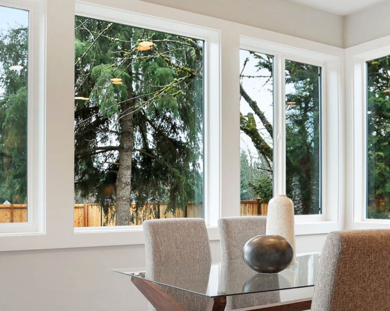 A bright dining area with large windows revealing pine trees outside. A glass table and upholstered chairs are present, with a decorative sphere centerpiece.